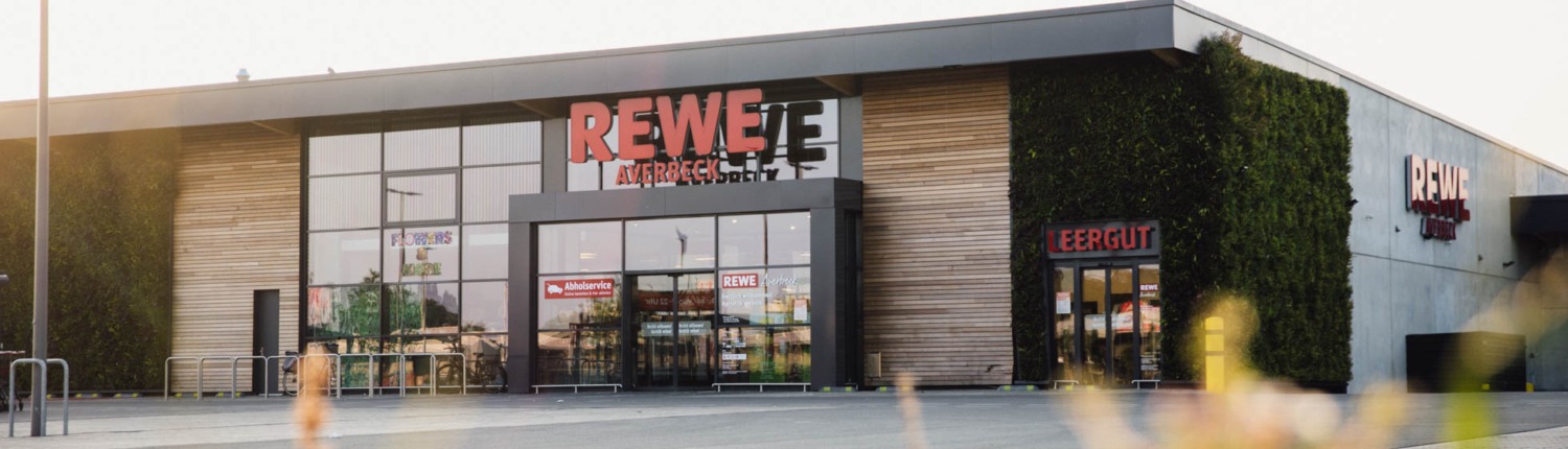 Moderne Außenansicht des REWE-Markts Averbeck in Kranenburg mit großen Glasfassaden, begrüntem Wandbereich und separatem Leergut-Eingang. Über dem Eingang steht der Text: ‚Kranenburger Unternehmen – Starke Partner in Kranenburg‘