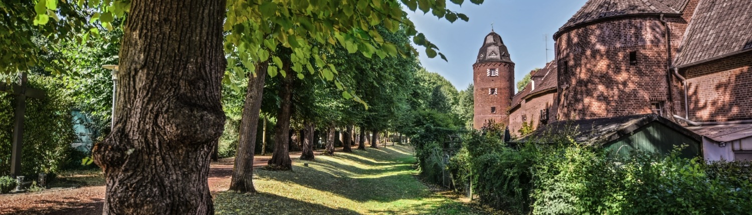 Baumgesäumter Fußweg mit Kopfsteinpflaster in Kranenburg, links große Laubbäume mit Schattenwurf, rechts historische Backsteinbauten und der markante Rundturm der Stadtmauer. In der Bildmitte steht in weißer Schrift: ‚Willkommen in Kranenburg – Ihr Tor zu Einkaufs- und Wohlfühlerlebnissen‘.