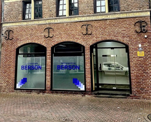 Straßenansicht der modernen Büroräume von Berson in Kranenburg, mit drei großen Fenstern in einer roten Backsteinfassade. Auf den Fenstern steht der Firmenname ‚BERSON‘ in blauer Schrift.