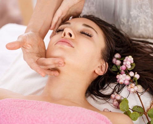 Entspannte junge Frau bei einer Gesichtsbehandlung, sie liegt mit geschlossenen Augen und wird von einer Dame im Gesicht masssiert