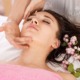 Entspannte junge Frau bei einer Gesichtsbehandlung, sie liegt mit geschlossenen Augen und wird von einer Dame im Gesicht masssiert