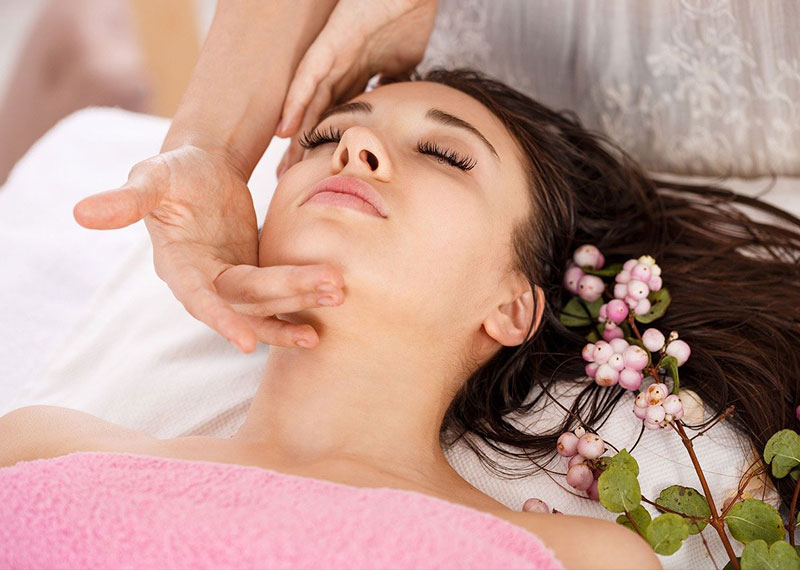 Entspannte junge Frau bei einer Gesichtsbehandlung, sie liegt mit geschlossenen Augen und wird von einer Dame im Gesicht masssiert