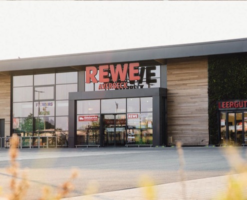 Moderne Außenansicht des REWE-Markts Averbeck in Kranenburg mit großen Glasfassaden, begrüntem Wandbereich und separatem Leergut-Eingang.