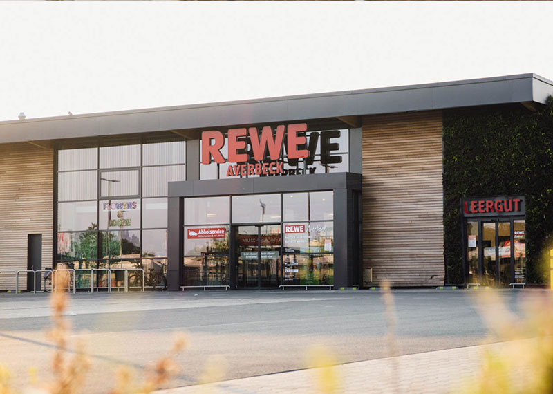 Moderne Außenansicht des REWE-Markts Averbeck in Kranenburg mit großen Glasfassaden, begrüntem Wandbereich und separatem Leergut-Eingang.