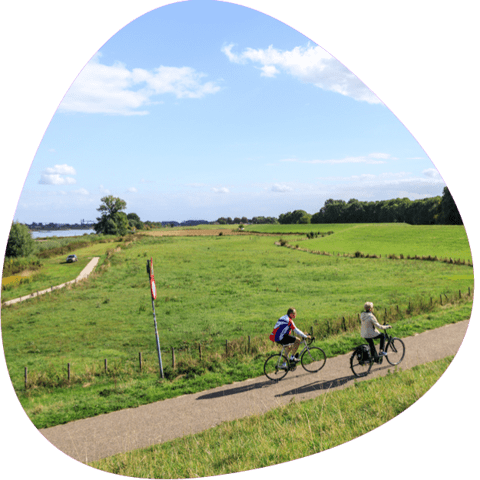 Zwei Radfahrer bei schönem Wetter vor grünen Wiesen mit einem Fluss im Hintergrund
