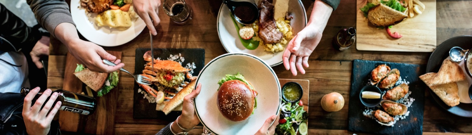 Fünf Personen am Essenstisch mit verschiedenen Gerichten (Hamburger, Steaks, Salate, Sandwiches etc.) Aufnahme von oben